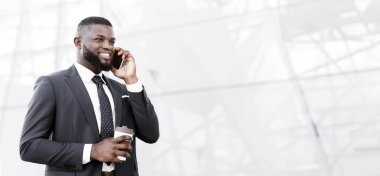 Gülümseyen Afro İş Adamı cep telefonuyla konuşuyor elinde kahve fincanıyla modern binanın yanında yürüyor. Boşluğu Kopyala