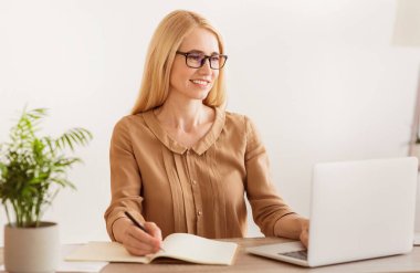 Bir kadın, dizüstü bilgisayarı üzerinde çalışırken bir not defterine not alıyor. Gülümsüyor ve odaklanmış görünüyor..