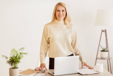 Gülümseyen bir kadın önünde dizüstü bilgisayarla bir masada duruyor. Açık renkli bir kazak ve siyah pantolon giyiyor..