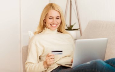Bir kadın elinde kredi kartıyla kanepeye oturmuş gülümsüyor ve internetten alışveriş için dizüstü bilgisayar kullanıyor..