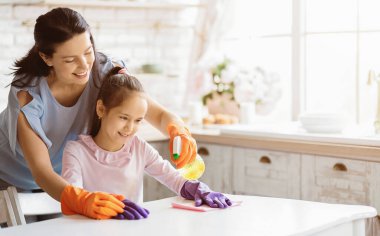 Bir anne ve kızı birlikte bir mutfak masasını temizliyorlar. Anne turuncu eldiven giyiyor ve kızı mor eldiven giyiyor. Birlikte çalışırken her ikisi de gülümsüyor, boşluğu kopyalayın.