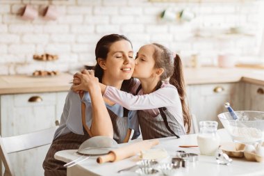 Genç bir kız, mutfakta birlikte kurabiye pişirirken annesini yanağından öpüyor. Annesinin yüzünde tatlı bir gülümseme var ve kızı ona sevgiyle bakıyor.