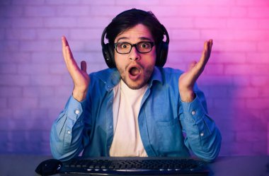 Bu resim, kulaklık takan, bilgisayarın önünde oturan ve şaşırmış ve şok olmuş bir genç adamı gösteriyor. Mavi bir gömlek giyiyor ve bir video oyunu oynuyor gibi görünüyor..