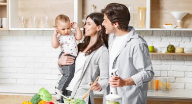 Üç kişilik genç bir aile mutfaklarında birlikte yemek hazırlıyor. Anne, bir kase salatayı karıştırırken ilgiyle bakan bebeklerini kucağında tutuyor..