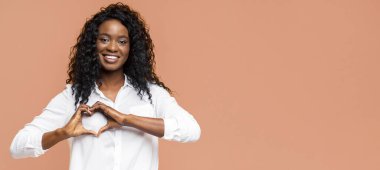 Kıvırcık siyah saçlı genç bir Afrikalı Amerikalı kadın gülümsüyor ve ellerini şeftali arka planda kalp şeklinde tutuyor. Kadın beyaz düğmeli bir gömlek giyiyor..