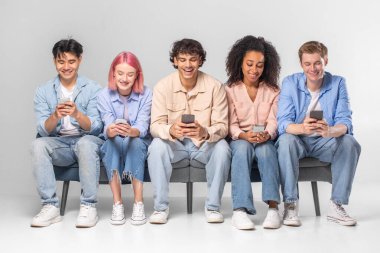Bu resim bir bankta birlikte oturan beş farklı genç yetişkini gösteriyor. Hepsi akıllı telefonlarına bakıyorlar. İnsanlar rahat ve rahat..