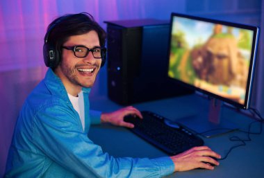 Mavi gömlekli ve kulaklıklı genç bir adam gece bilgisayarda bir video oyunu oynuyor. Gülümsüyor ve iyi vakit geçiriyor gibi görünüyor. Bilgisayar ekranı oyunun renkleri ile aydınlanır