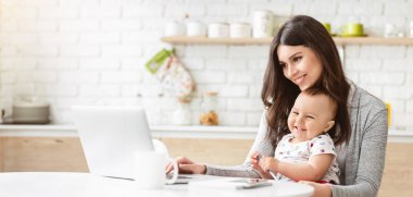 Mutfakta bir masada oturan bir kadın dizüstü bilgisayarla çalışıyor. Gülümsüyor ve kucağında bir bebek var. O da gülümsüyor ve kameraya bakıyor..