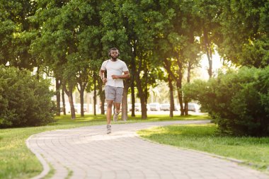 Beyaz tişörtlü ve gri şortlu bir adam parktaki kaldırım yolunda koşuyor. Güneşli bir yaz gününün tadını çıkarırken müzik dinliyor..