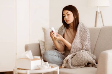 Genç bir kadın battaniyeyle kaplı bir kanepede oturur ve bir şişe ilaca bakar..