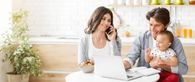 Kocası yanında bebeğini tutarken bir kadın mutfak masasında dizüstü bilgisayarla oturup telefonla konuşuyor. Bebek dizüstü bilgisayara bakıyor ve kadın gülümsüyor..