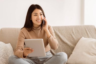 Kanepede oturan genç bir kadın elinde tabletle telefonla konuşurken gülümsüyor..