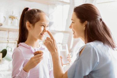 Bir anne ve kızı mutfakta neşeli bir anın tadını çıkarıyorlar. Kız elinde bir kek tutuyor ve gülümsüyor. Bu arada anne de burnuna dokunuyor..