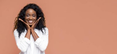 Kıvırcık saçlı, gülümseyen siyah bir kadın şeftali renkli bir arka planın önünde elleri yanaklarında poz veriyor. Beyaz düğmeli bir gömlek giyiyor..