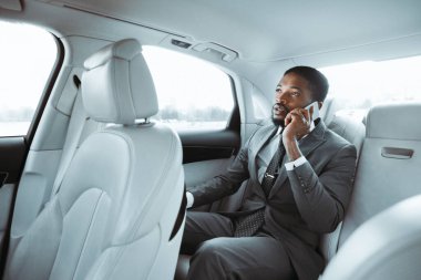 Gri takım elbiseli Afro-Amerikalı işadamı bir arabanın arka koltuğunda oturuyor, telefonuyla konuşurken pencereden dışarı bakıyor. Arabanın içi açık renkli deri..