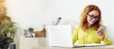 Kızıl saçlı ve gözlüklü bir kadın akıllı telefonuna bakarken gülümsüyor. Elinde dizüstü bilgisayar ve not defteriyle bir masada oturuyor..