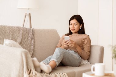 Genç bir kadın bej bir kanepede dinleniyor, bacaklarını açıyor ve telefonuna bakıyor..