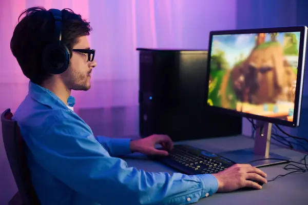 stock image A young man wearing headphones is playing a computer game at a desk in his home office. He is focused on the screen and is using a keyboard and mouse to control the game.