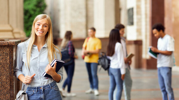Счастливая студентка с книгами, позирующими перед камерой в студенческом зале, с перерывом между занятиями
