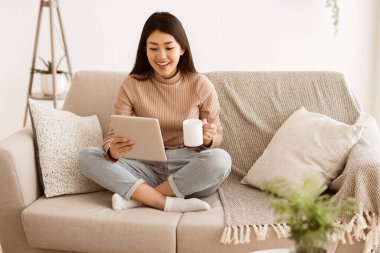 Bir kadın elinde tablet ve bir fincan kahve tutarken kanepede dinlenir. O, gündelik giysiler giyer ve yüzünde rahat bir ifade vardır..