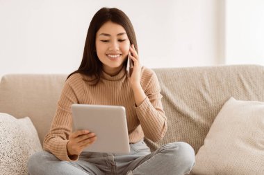 Bir kadın kanepede oturuyor, tablet kullanıyor ve telefonda konuşuyor..