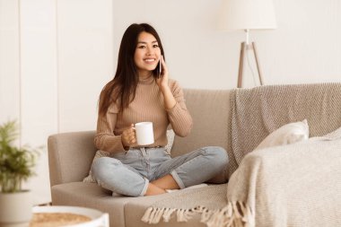 Bir kadın bir kanepede oturuyor, elinde bir kupa kahve tutarken gülümsüyor ve telefonuyla konuşuyor..