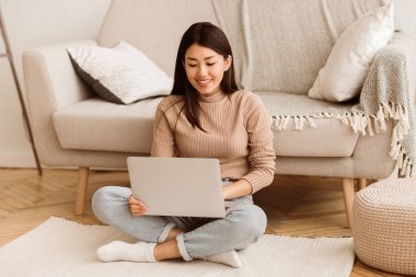 Genç bir kadın oturma odasında yerde oturuyor, gülümserken dizüstü bilgisayar kullanıyor..