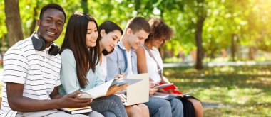 Smiling African American Student Guy Studying Outdoors With His University Friends, Holding Smartphone And Smiling At Camera, Selective Focus