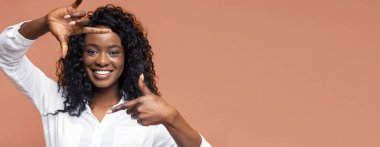 Görüntü, siyah kıvırcık saçlı Afro-Amerikalı bir kadını, elleri şeftali rengi bir arka planda çerçeve yaparken gülümserken gösteriyor. O beyaz bir gömlek giyiyor..
