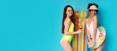 Bu görüntüde iki genç kadın mayo giyiyor ve şişme havuz ile parlak mavi bir arka plana karşı poz veriyor.