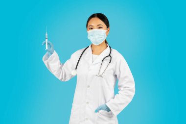 Beyaz önlük, yüz maskesi ve mavi eldiven giyen Asyalı kadın doktor sağ elinde bir şırınga tutuyor. Mavi bir arka planda duruyor.