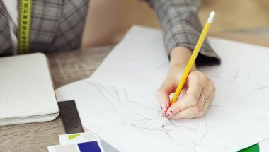 Bir moda tasarımcısı sarı bir kalem kullanarak beyaz bir kağıt üzerine bir elbise tasarımı çizer..