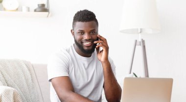 Gülümseyen siyah bir adam akıllı telefonuyla konuşurken evdeki bir kanepede oturuyor. Beyaz bir tişört giyiyor ve önünde bir laptop var..