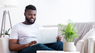 Afro-Amerikalı bir adam oturma odasındaki açık gri bir kanepede oturuyor, gülümserken dizüstü bilgisayar kullanıyor. Arka planda bir saksı bitkisi ve yer lambası görünür..