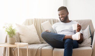 Siyah adam bir elinde tablet, diğer elinde kahve fincanı tutarken bacakları havada bir kanepede oturuyor, gülümsüyor. Kanepe ışıl ışıl aydınlatılmış bir oturma odasında.