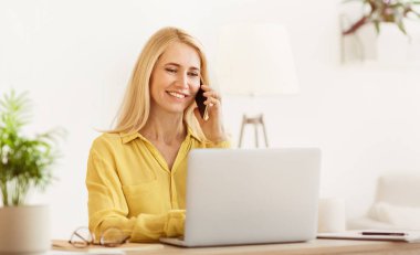 Gülümseyen bir kadın, arka planda bir bitkiyle telefonuyla sohbet ederken parlak ev bilgisayarını tamir ediyor..