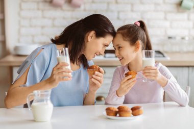 Bir anne ve küçük kızı ellerinde süt ve muffin bardaklarıyla mutfak masasında oturuyorlar. Birbirlerine gülümsüyorlar ve birlikte mutlu bir anın tadını çıkarıyor gibi görünüyorlar.