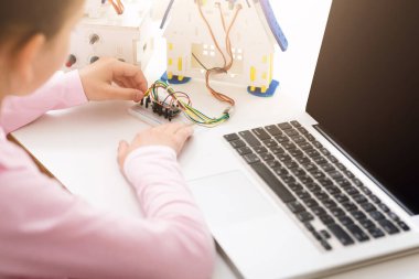 Kız bir masada elektronik bir proje üzerinde çalışıyor, kabloları ekmek tahtasına bağlıyor. Masada küçük bir model ev ve dizüstü bilgisayar da bulunuyor, bu da projenin robotikle ilgili olduğunu gösteriyor..