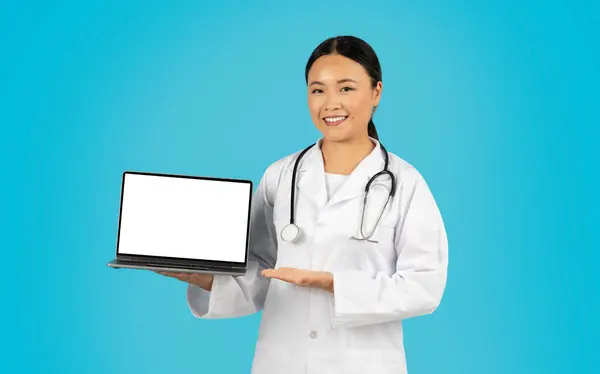 stock image Asian woman doctor wearing a white coat and stethoscope smiles and holds a laptop with a blank screen in front of her. The background is a solid blue color.