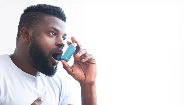 stock image An image of African American man using an inhaler to treat his asthma. He is holding the inhaler in his right hand and has his mouth open. He is wearing a white t-shirt, copy space