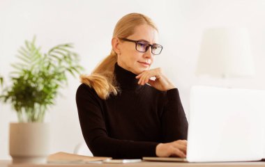 Gözlüklü bir kadın iyi aydınlatılmış bir ofiste dizüstü bilgisayarıyla çalışıyor..