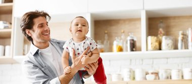 Gülümseyen bir baba küçük oğlunu mutfakta tutuyor. Küçük çocuk kameraya tuhaf bir ifadeyle bakıyor. Mutfak modern ve iyi ışıklandırılmış, beyaz dolaplar ve seramik kaplama.