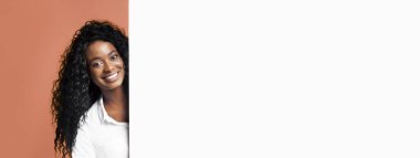 Uzun kıvırcık siyah saçlı genç bir kadın beyaz bir duvarın arkasından bakarken kameraya gülümsüyor. Beyaz düğmeli bir gömlek giyiyor ve arkasında şeftali rengi bir arka plan var..