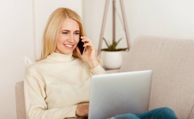 Bir kadın kanepede oturuyor, cep telefonuyla konuşurken gülümsüyor ve laptopuyla çalışıyor..