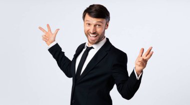 Siyah takım elbiseli bir işadamı iki eliyle beyaz bir arka plana gülümser ve el kol hareketi yapar..