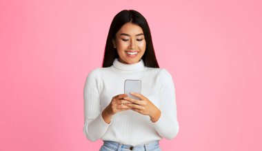 Asyalı genç bir kadın akıllı telefonunu kullanırken pembe bir arka planın önünde duruyor. Gülümsemesi, telefonda gördüklerinin hoşuna gittiğini gösteriyor..