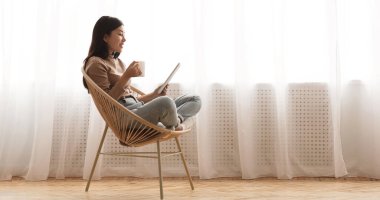 Boş zaman. Tablet kullanan ve kahve içen bir kız, pencereye karşı hasır sandalyede oturan, boş alanı olan bir panorama