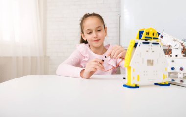 Beyaz bir masada oturan genç bir kız robot setini birleştirmeye odaklanmış. Elinde küçük bir kablo tutuyor, robotların kablolarına dikkatlice bağlıyor. Robot beyaz plastikten yapılmıştır.