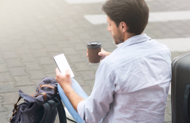 Mekik Otobüsü Bekliyor. Genç adam kahve içiyor ve akıllı telefon kullanıyor havaalanı binasının yanında otururken, Omuz manzaralı, bedava uzay manzaralı.