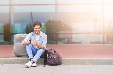 Uçuşlar Arası Yedek Zaman. Millennial Guy, bavul ve akıllı telefonlu, açık havada dinlenen, boş alanı olan havaalanı yakınında oturuyor.